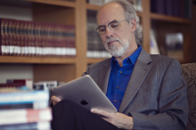 Image of a man with grey hair, glasses, goatee wearing a blue shirt with a grey jacket. Jamie is holding a Mac laptop and sitting in front of a bookshelf filled with books.