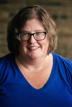 Image of a woman with brown hair, black glasses, and wearing a blue shirt in front of a blurred background.