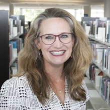 Image of a woman with light brown hair, red glasses, wearing a white and black shirt. The photo is in front of library stacks.