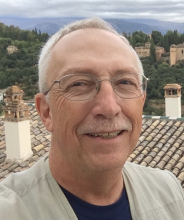 Image of man wearing a cream colored sweater over a blue shirt, with wire-framed glasses, white hair standing in front of a landscape.